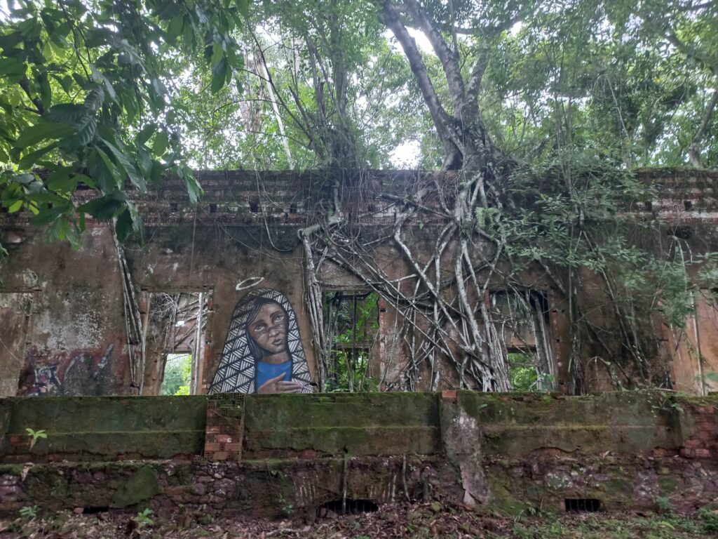 Eine Mauer mit mehreren Durchgängen. Neben dem linken Durchgang befindet sich ein guterhaltenes, mannsgroßes Mosaik eines Heiligenbildes, die anderen Zwischenmauern werden von Luftwurzel der auf der Mauer wachsenden Bäume bedeckt.